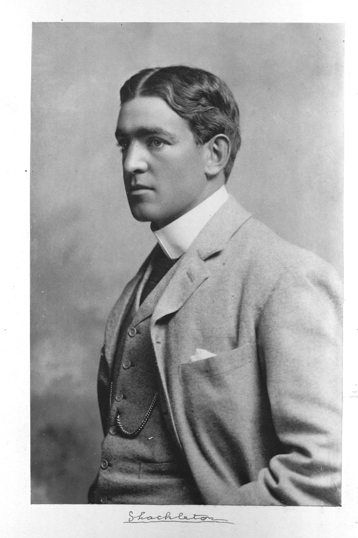 Black and white portrait of Ernest Shackleton. Side view. Shackleton is wearing a light grey suit jacket and white collared shirt. Plain backdrop.